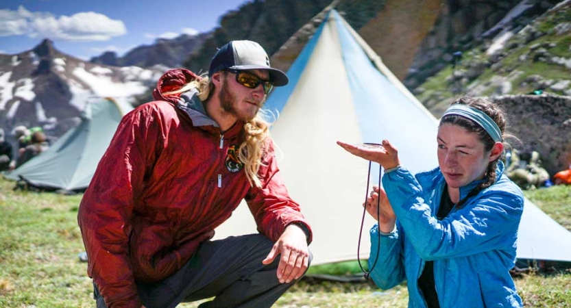 Two people examine a compass while sitting in front of a tent in an alpine landscape